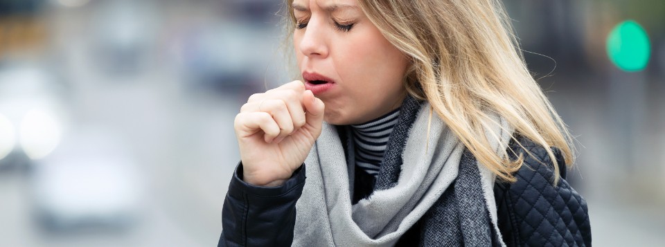 Woman coughing outdoors while wearing a scarf and winter coat, experiencing symptoms of a persistent cough.